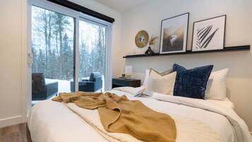 Une chambre moderne avec un grand lit, une table de chevet, une fenêtre donnant sur la forêt et des œuvres d’art encadrées accrochées au mur.