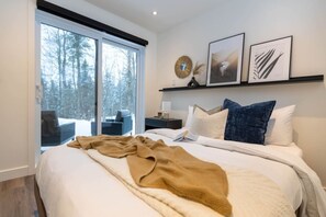 A modern bedroom with a large bed, a nightstand, a window with a view of trees, and framed artwork on the wall. - Private Spa| Hiking| Skiing| Mini Chalet| Assinie (Brownsburg)