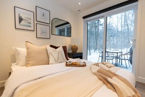 A hotel room with a large bed, a bedside table with a lamp, a tray with breakfast items, and a view of a snowy outdoor area through a sliding door. - Private Spa| Hiking| Skiing| Mini Chalet| Assinie (Brownsburg)