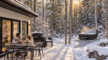 Et udendørs spiseområde dækket af sne med jacuzzi og grill, omgivet af skov.