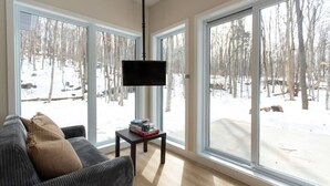 A living room with a sofa, a coffee table, and a TV. Large windows offer a view of a snowy outdoor area. - Private Spa | Hiking | Skiing | Mini - Prime Hill (Brownsburg)
