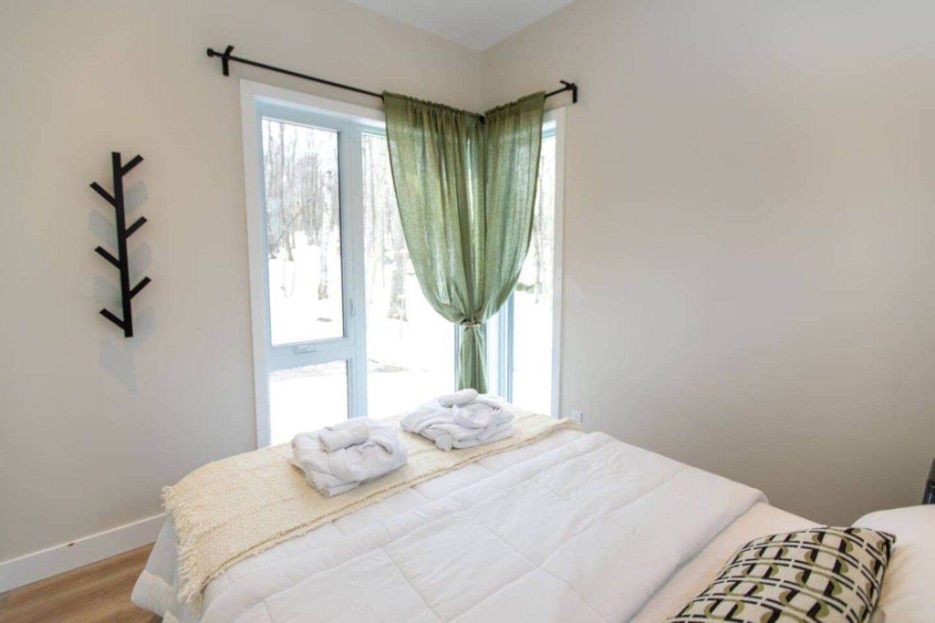 A bedroom with a bed, a window with curtains, a wall-mounted coat rack, and a view of trees outside.