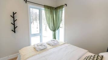 A bedroom with a bed, a window with curtains, a wall-mounted coat rack, and a view of trees outside.