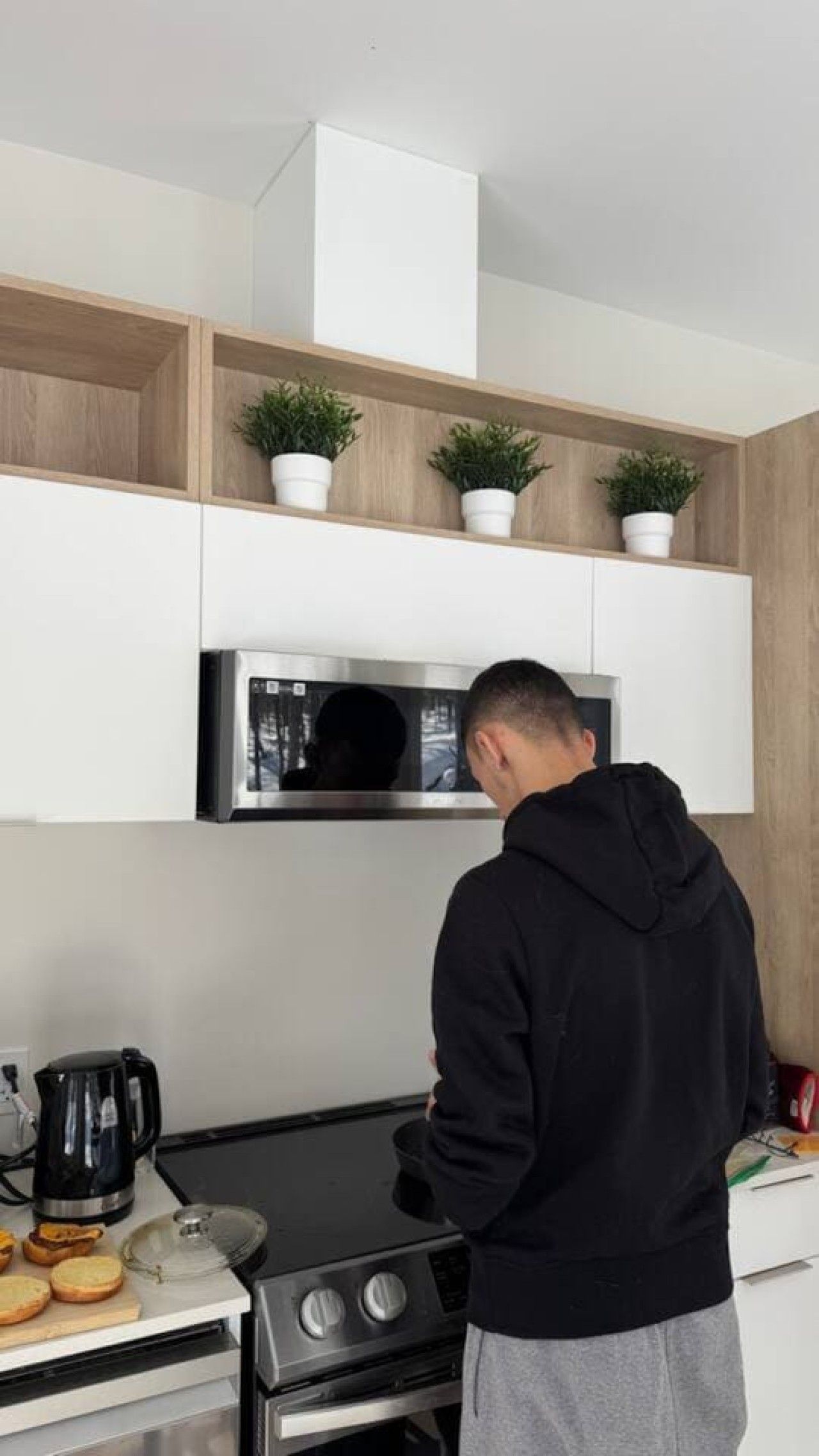 A person in a black hoodie cooking on a stove in a kitchen with white cabinets, a microwave, and potted plants on the shelves.