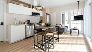 A modern kitchen with white cabinetry, a wooden dining table, and a bar stool. There is a large window providing natural light and a view outside. - Private Spa | Hiking | Skiing | Mini - Prime Hill (Brownsburg)
