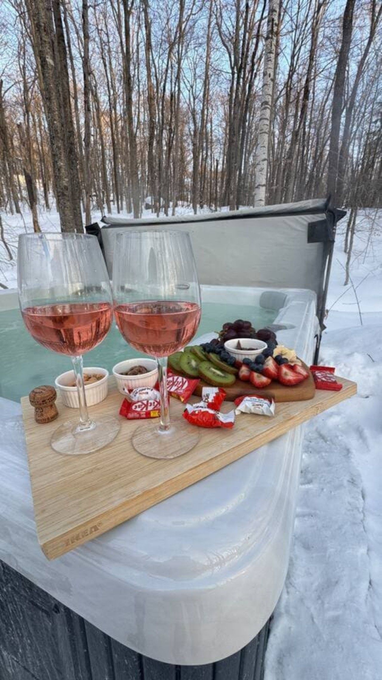 Un montaje para picnic con dos copas de vino, una tabla de madera con frutas y aperitivos, y una tienda de campaña en un bosque nevado.