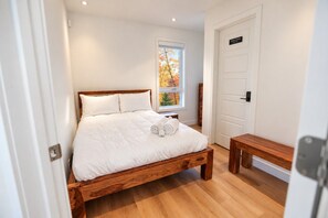 Un dormitorio con cama de madera, ropa de cama blanca, un banco, un ventanal con vistas a árboles otoñales y una puerta que indica 'Solo personal'.