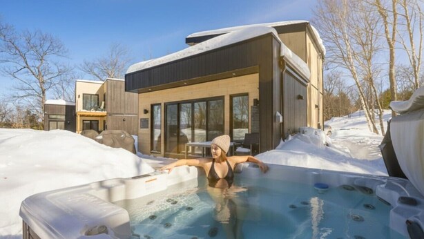 A person relaxing in a hot tub outside a modern house with large windows and a covered patio.