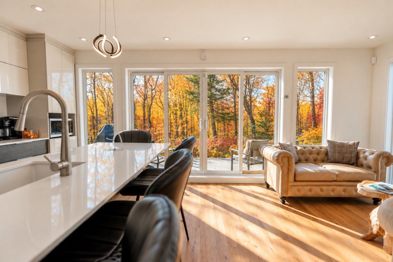 A modern kitchen with a central island, dining chairs, and a sofa facing a large sliding glass door with a view of autumn trees.