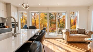 A modern kitchen with a central island, dining chairs, and a sofa facing a large sliding glass door with a view of autumn trees.