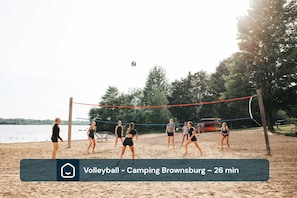 A beach volleyball game with players in sportswear, a volleyball net, and a sandy beach with trees in the background.
