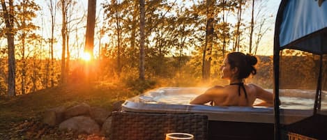 A person relaxing in a hot tub with a view of trees and the sun setting in the background.