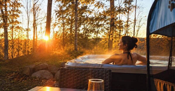 A person relaxing in a hot tub with a view of trees and the sun setting in the background. - Private Spa| Hiking | Skiing | Mini Chalet Élios (Argenteuil Regional County Municipality)