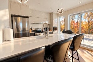 Una cocina moderna con una isla grande, electrodomésticos de acero inoxidable y vistas a árboles otoñales.