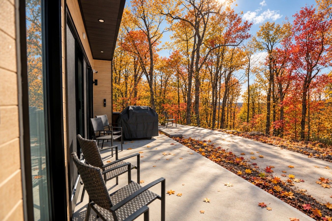 Un espace extérieur aménagé, abrité et doté de chaises et d’un barbecue, offrant une vue sur une forêt aux couleurs automnales.