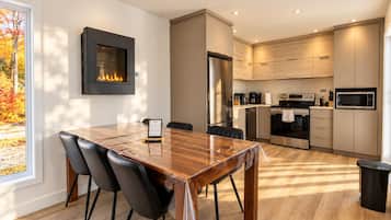 A modern kitchen with a dining area, a fireplace, and a view of autumn foliage.