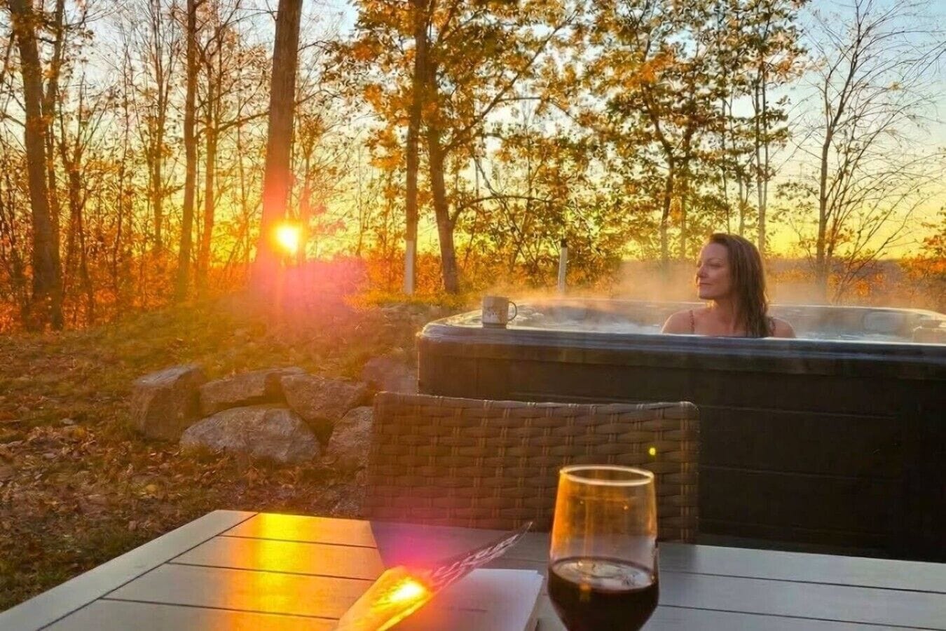 A person relaxing in a hot tub with a glass of wine and a candle on a table in a forested area.