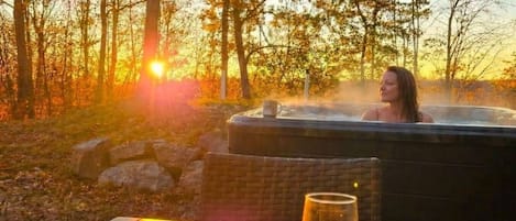 Uma pessoa relaxando em uma banheira de hidromassagem com uma taça de vinho e uma vela sobre uma mesa em uma área arborizada.