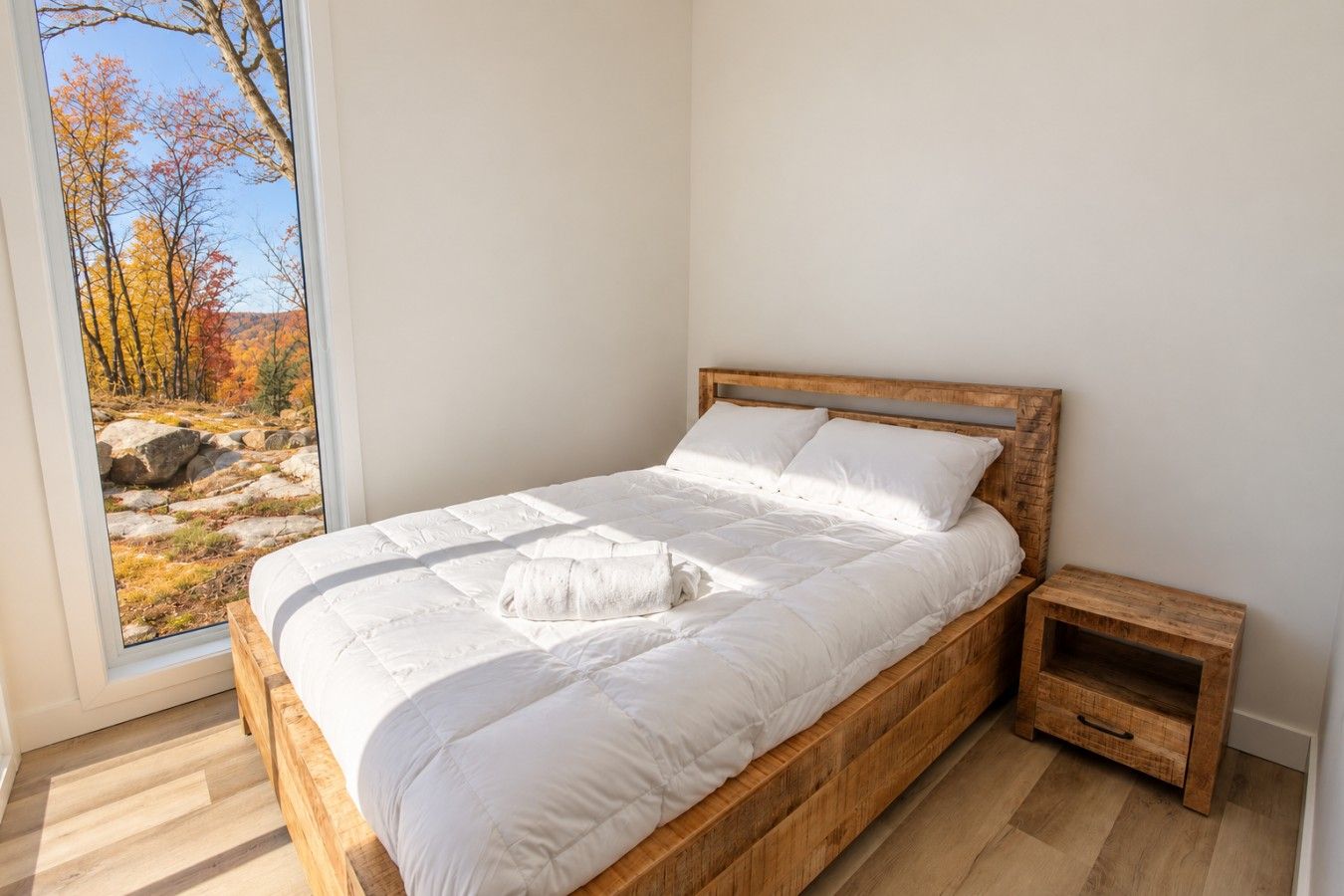 A bedroom with a wooden bed, white bedding, a nightstand, and a view of autumn trees through a window.