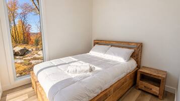 A bedroom with a wooden bed, white bedding, a nightstand, and a view of autumn trees through a window.