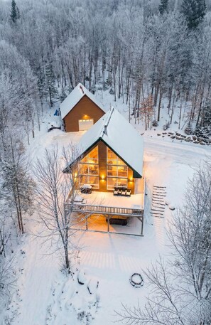 A cabin with a large front porch and a garage, surrounded by a snowy forest. - Private Hot Tub | Hiking | Skiing | Carriole (Val-des-Lacs)