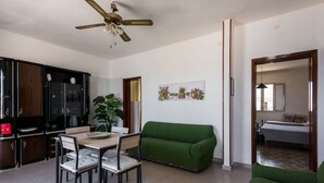 A modern living room with a green sofa, a dining table with chairs, a ceiling fan, and a kitchen area with cabinets and a plant. - La Casa Dell Isola by Wonderful Italy (Syracuse)