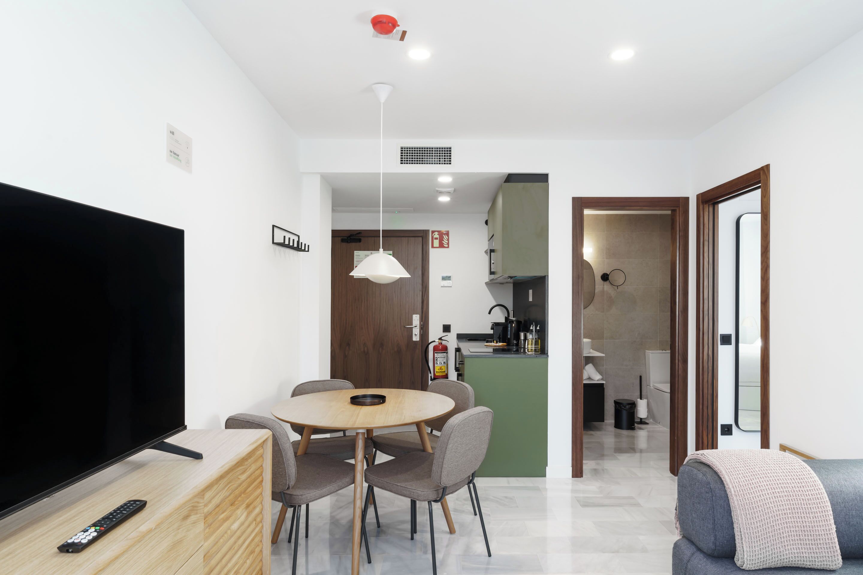 A modern living room with a flat-screen TV, a dining table with chairs, a sofa, and a kitchen area in the background.