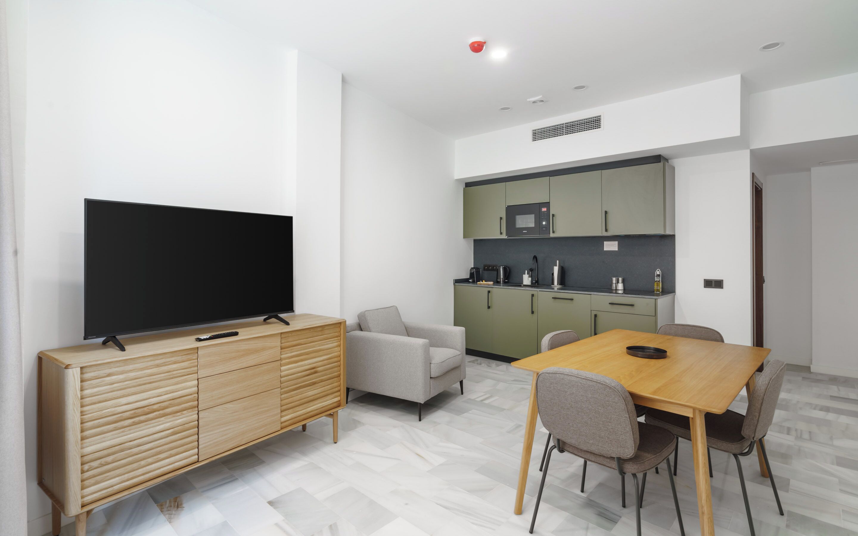 A modern living room with a flat-screen TV, wooden cabinet, grey sofa, and dining table with chairs.