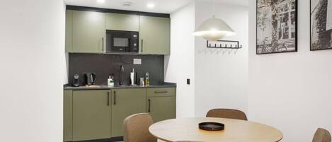 A modern kitchen with green cabinets, a microwave, and a coffee maker. There is a round table with chairs and a pendant light above it. Two framed pictures are on the wall.