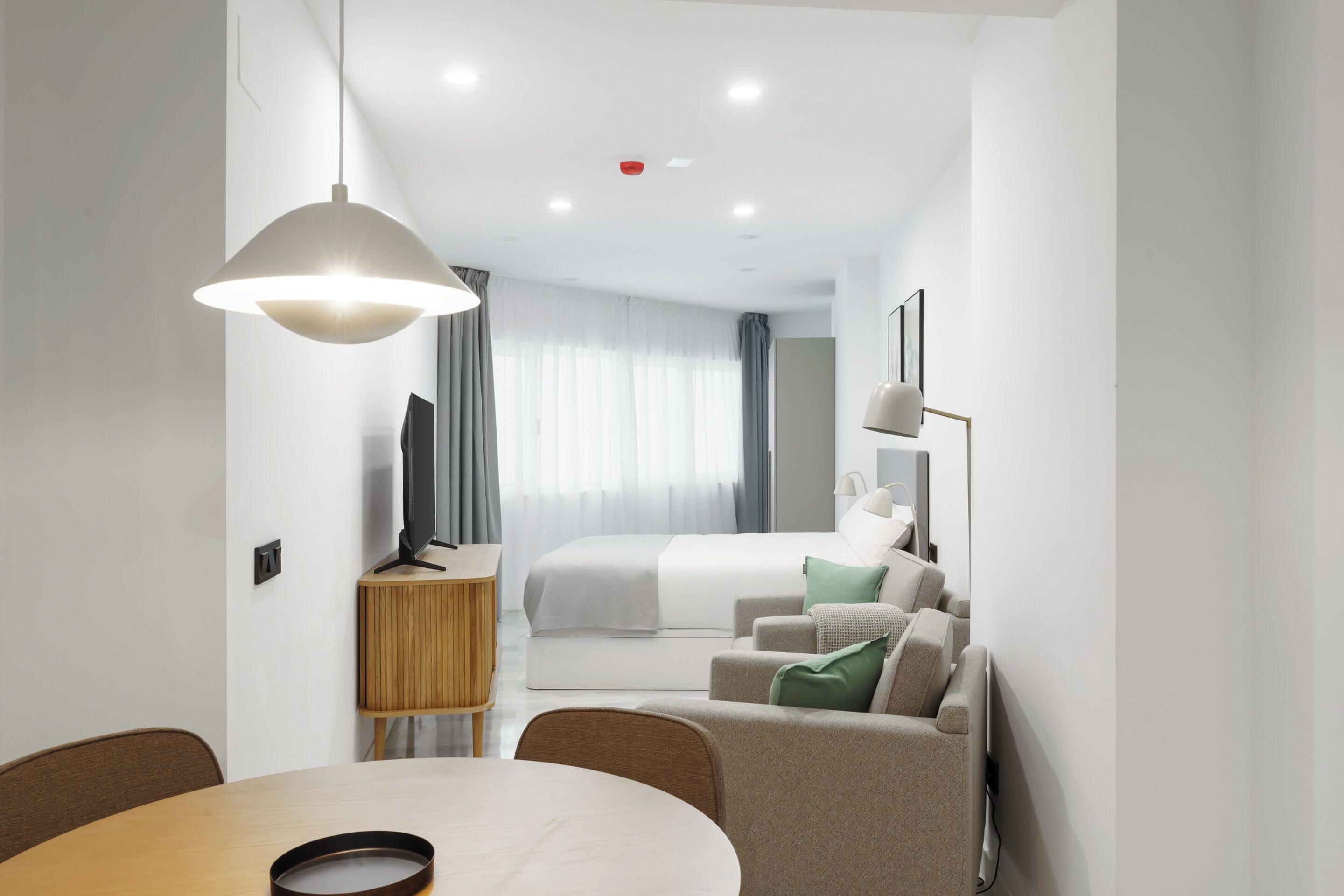 A modern hotel room with a bed, a sofa, a TV, a dining table, and a pendant light.