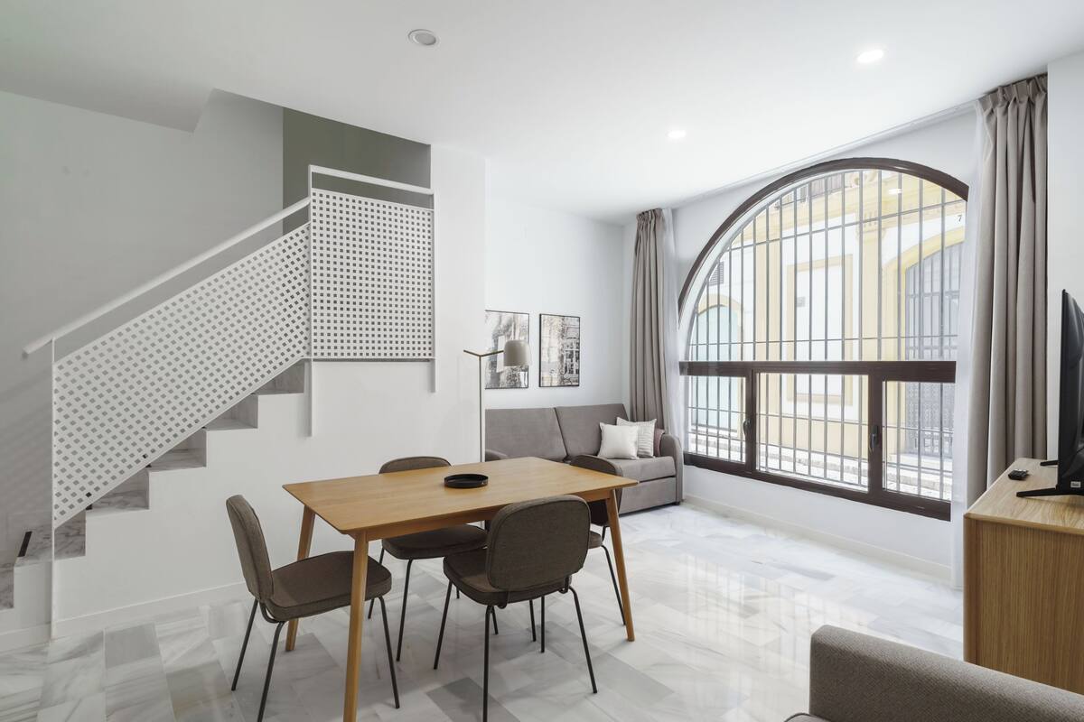 Una sala moderna con comedor, una escalera con barandilla estampada y un ventanal con cortinas.