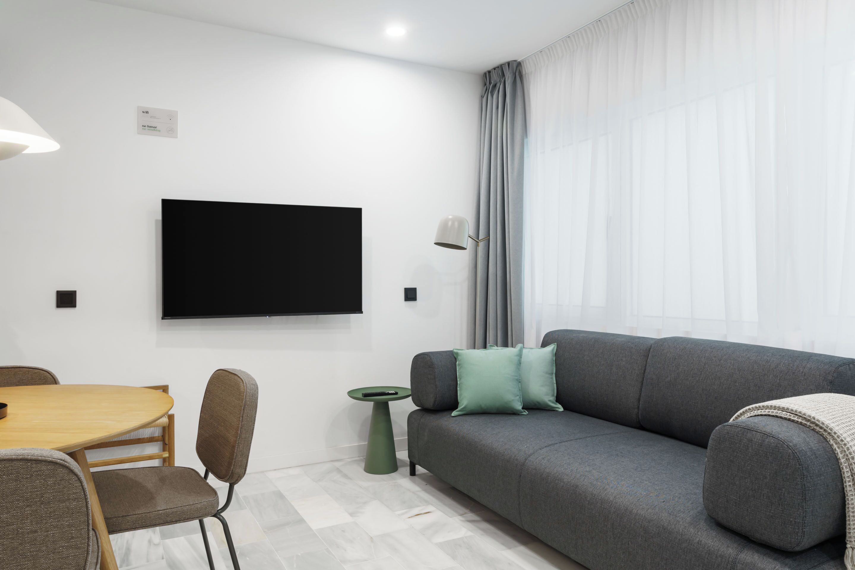 Ein modernes Wohnzimmer mit flachbildfernseher an der Wand, einem grauen Sofa mit Kissen, einem Holzesstisch mit Stühlen und einem kleinen Beistelltisch.