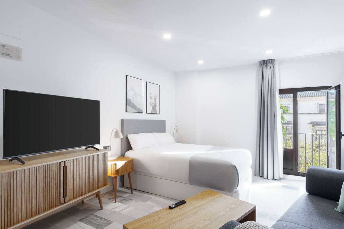 Una habitación de hotel moderna con una cama grande, un televisor sobre una consola de madera, una mesita y un sofá.