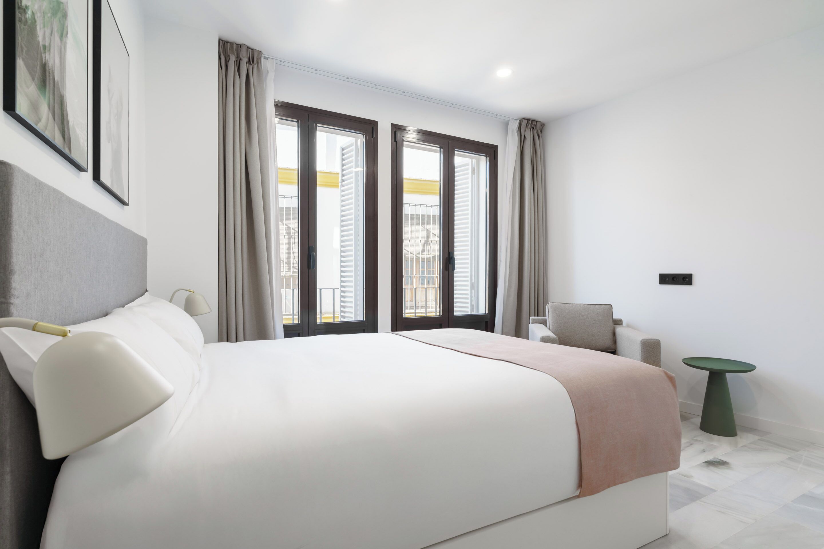 A modern hotel room with a large bed, two windows with curtains, a small table, and a chair.