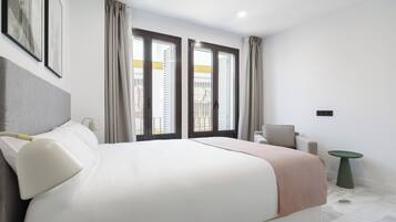 A modern hotel room with a large bed, two windows with curtains, a small table, and a chair.