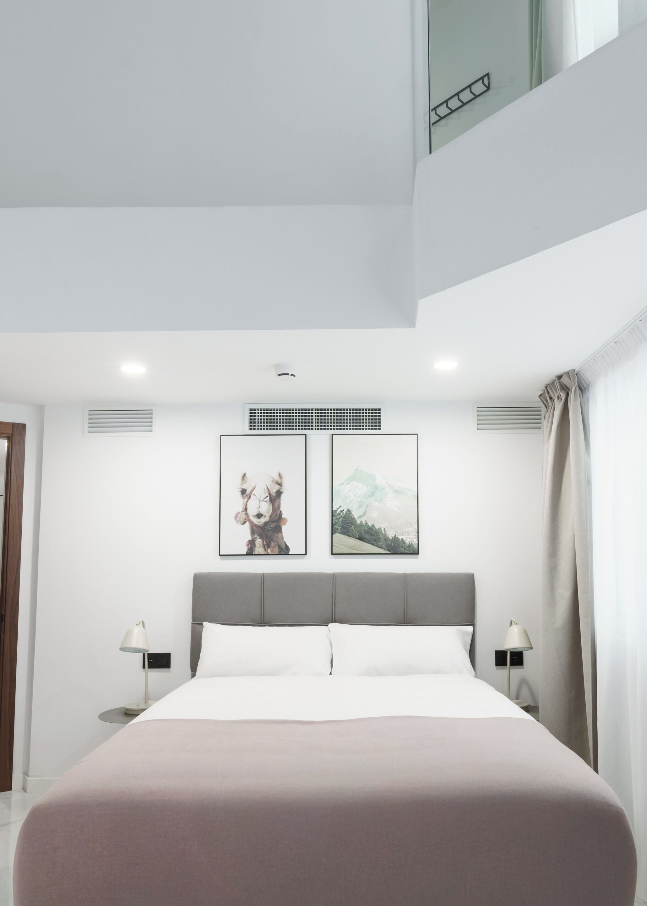 Un dormitorio moderno con una cama grande, dos mesitas de noche con lámparas, un cabecero gris y dos cuadros enmarcados en la pared.