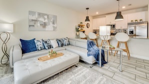 Un salon moderne avec un canapé d’angle blanc, une table basse en verre et un coin repas comprenant des chaises blanches et une petite table.