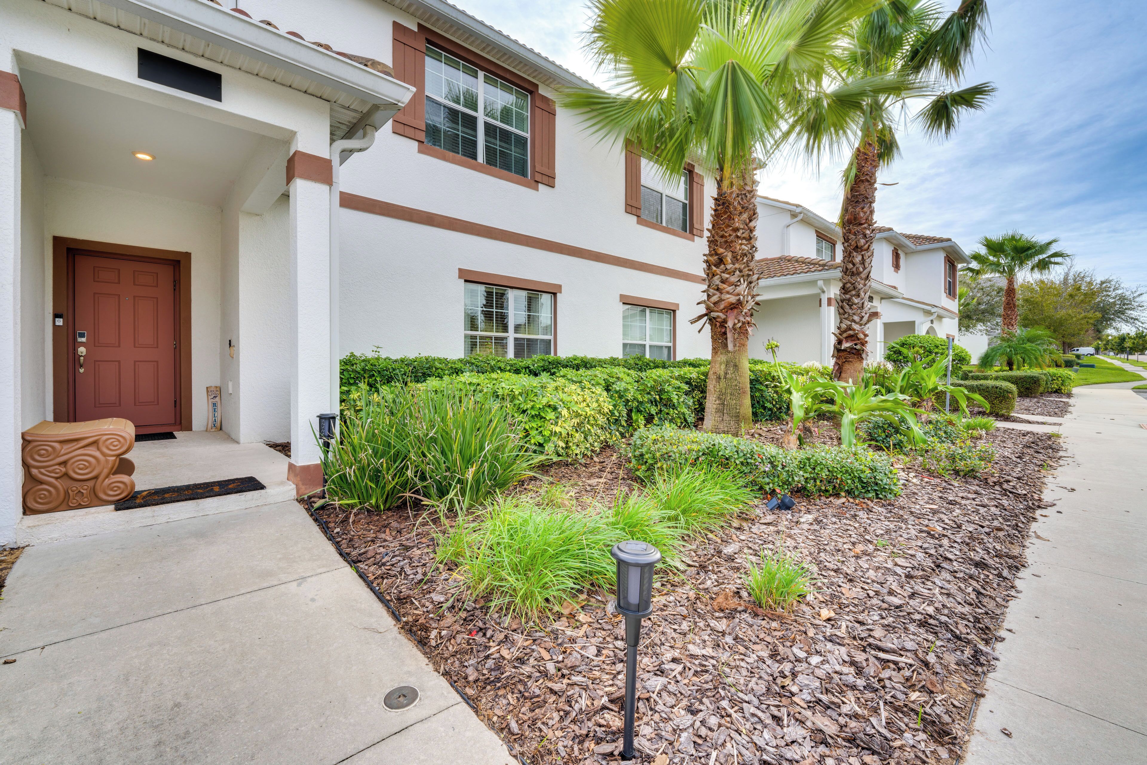 A residential building with a front entrance, a well-maintained garden with palm trees and shrubs, and a clear sky.