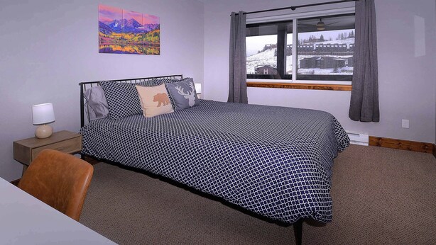 A bedroom with a bed, a nightstand, a lamp, a chair, a window with curtains, and a painting on the wall. - The Evergreen. Hot tub, grill, walk to slopes. (Crested Butte)