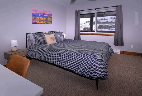 A bedroom with a bed, a nightstand, a lamp, a chair, a window with curtains, and a painting on the wall.