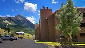 A building with a parking lot, a mountain in the background, and a tree in the foreground. - The Evergreen. Hot tub, grill, walk to slopes. (Crested Butte)