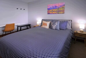 A neatly arranged bedroom with a patterned bedspread, a chair, a bedside table, and a wall-mounted painting.