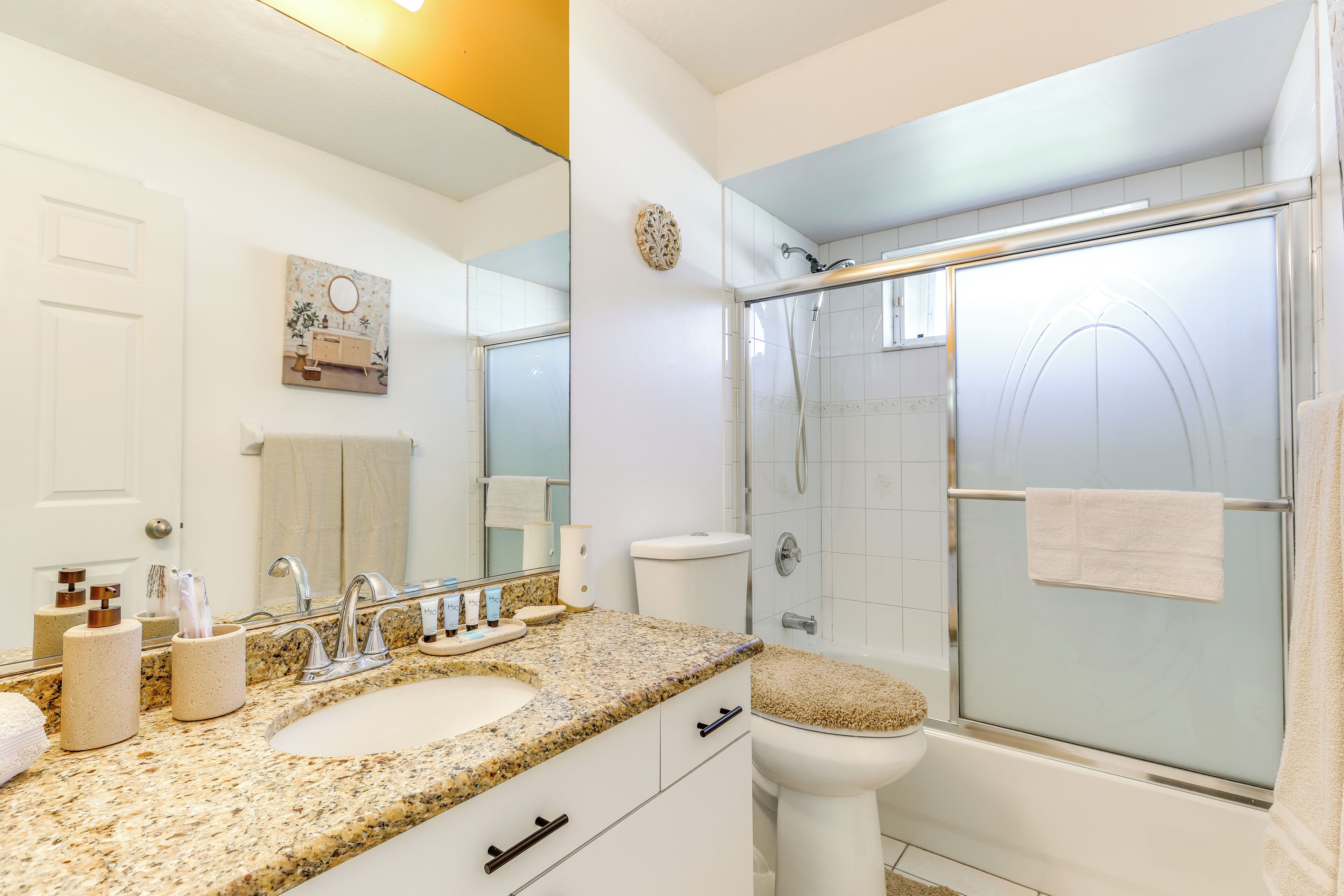 A bathroom with a granite countertop sink, a toilet, a glass-enclosed shower, and a mirror with a decorative wall piece.