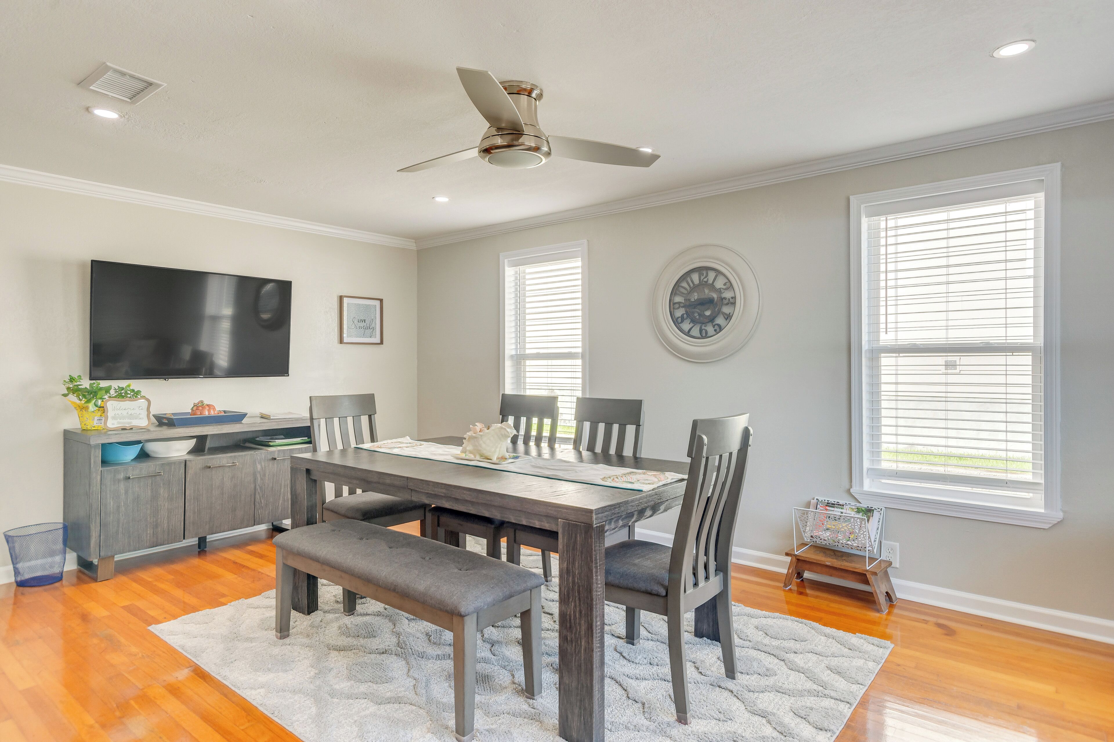 A dining area with a wooden table, chairs, and a bench. There is a ceiling fan, a clock on the wall, and a window with blinds.