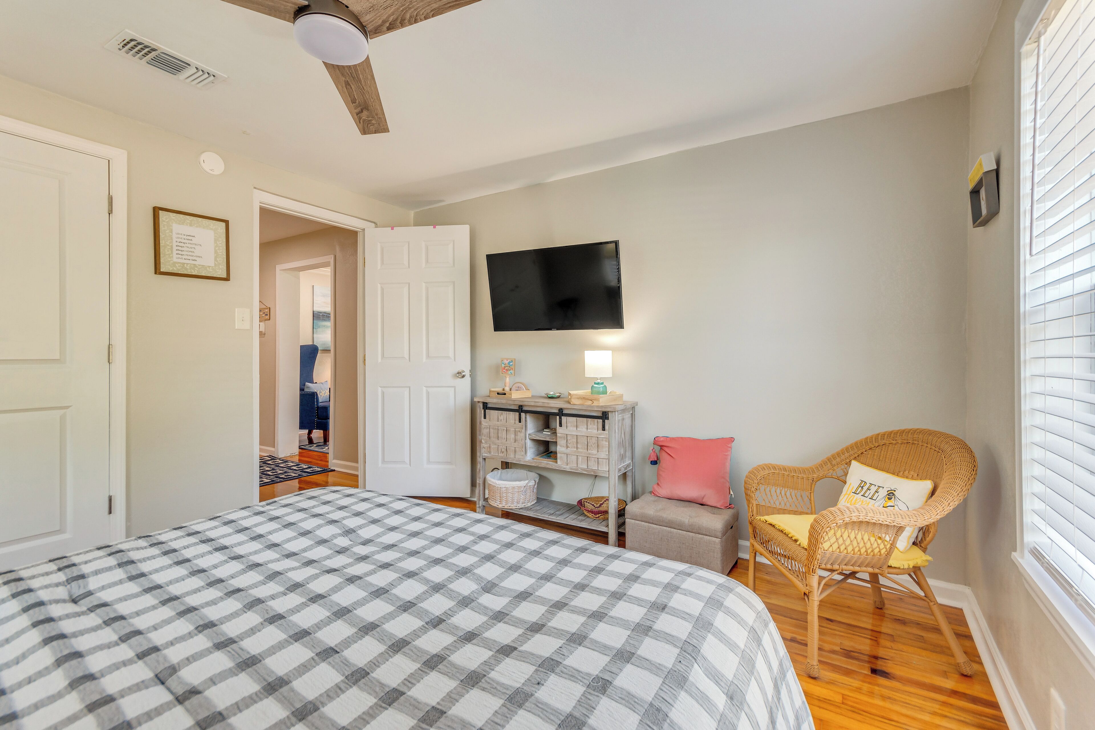 A bedroom with a checkered bedspread, a wicker chair with a cushion, a small table with a lamp, and a television on the wall.
