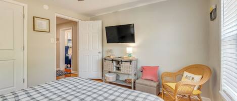 A bedroom with a checkered bedspread, a wicker chair with a cushion, a small table with a lamp, and a television on the wall.