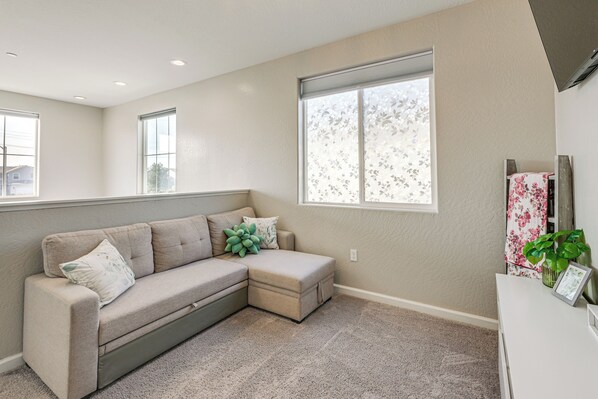 A living room with a sectional sofa, a small ottoman, a white console table with a plant, and a window with a floral view.