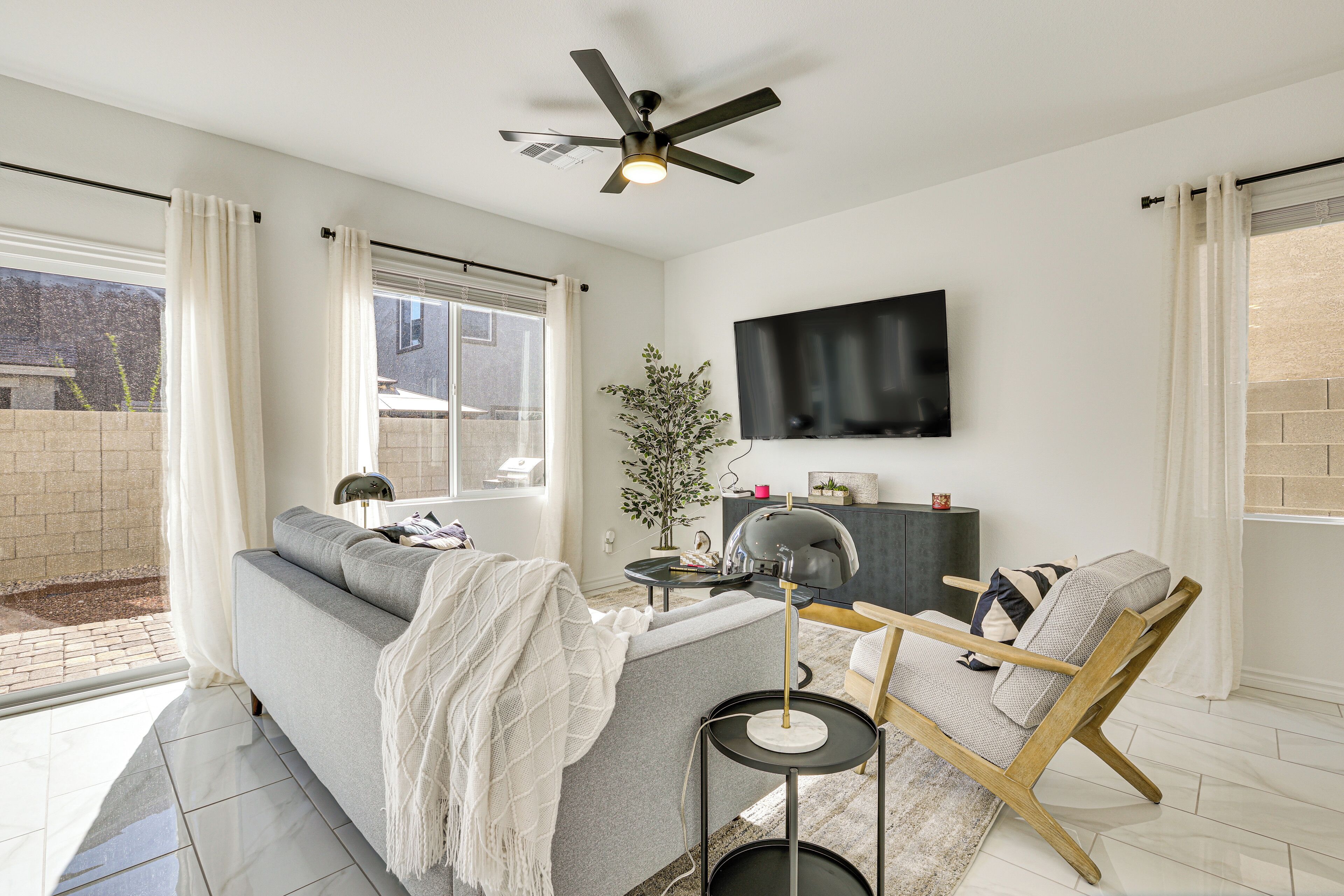 A modern living room with a grey sofa, a wooden chair, a ceiling fan, a TV mounted on the wall, and a potted plant.