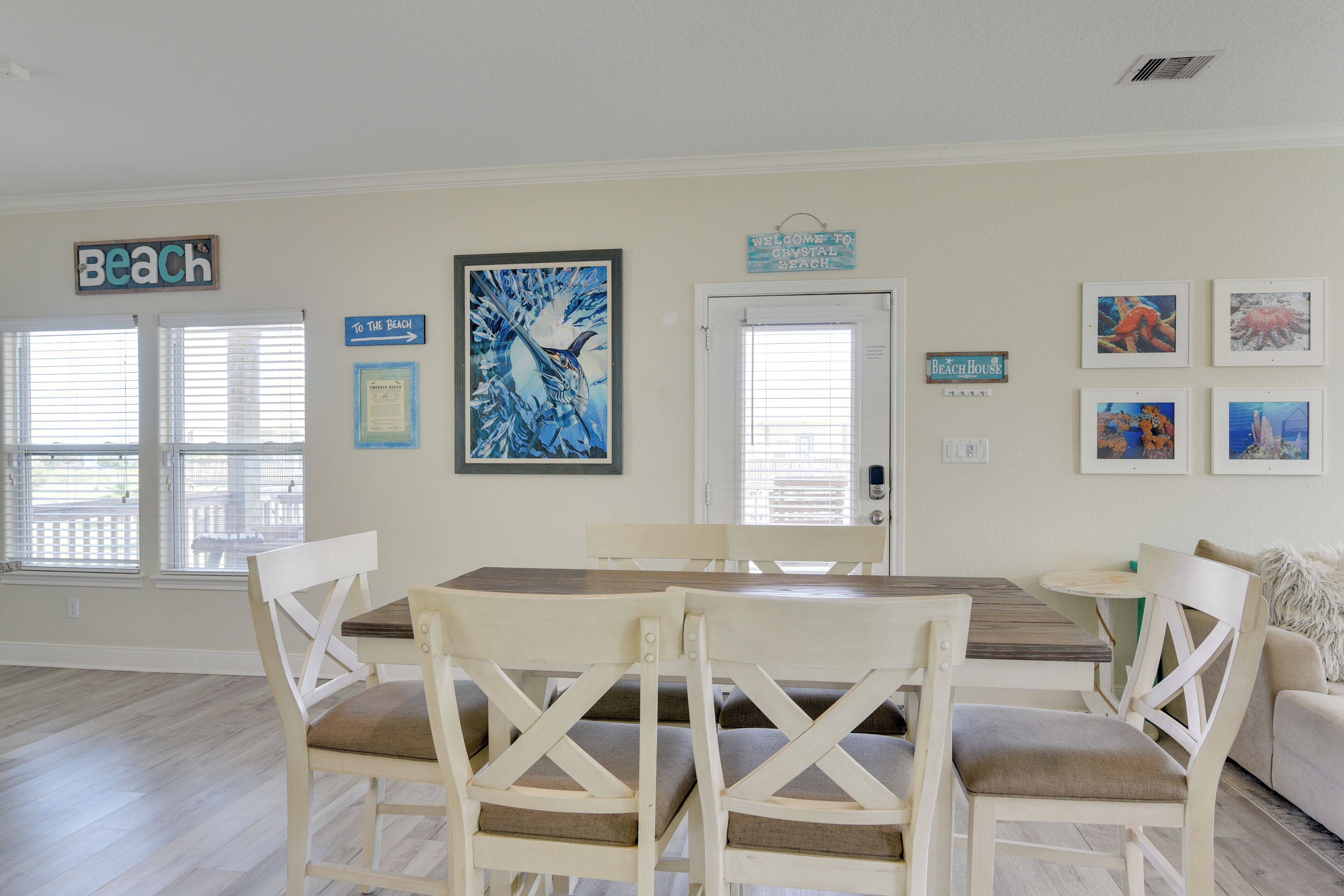 Un coin repas comprenant une table et des chaises, une porte et des éléments de décoration murale, notamment un panneau indiquant « Plage » et des tableaux encadrés.