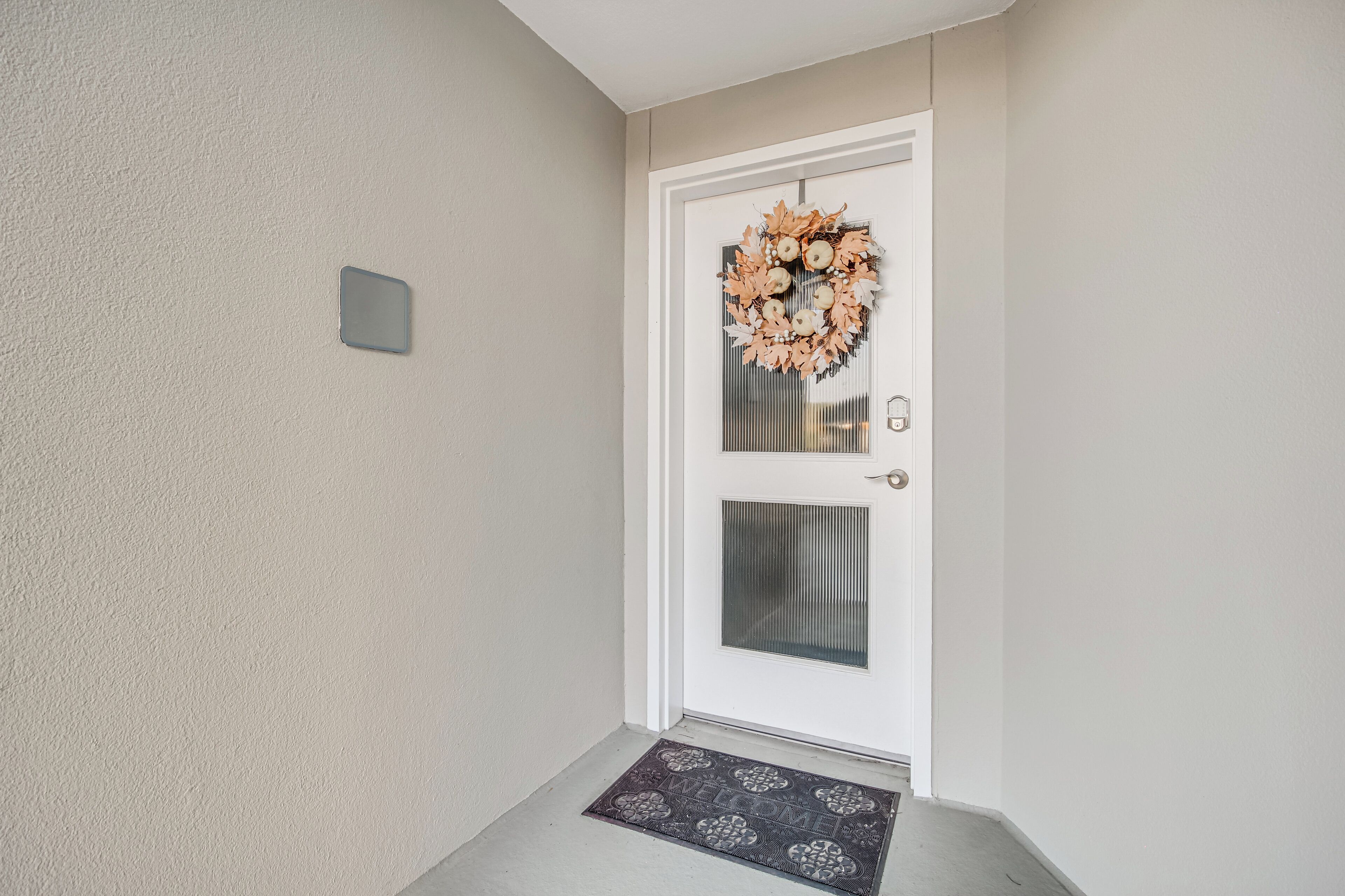 A white door with a decorative wreath, a small rectangular light switch, and a welcome mat at the entrance.